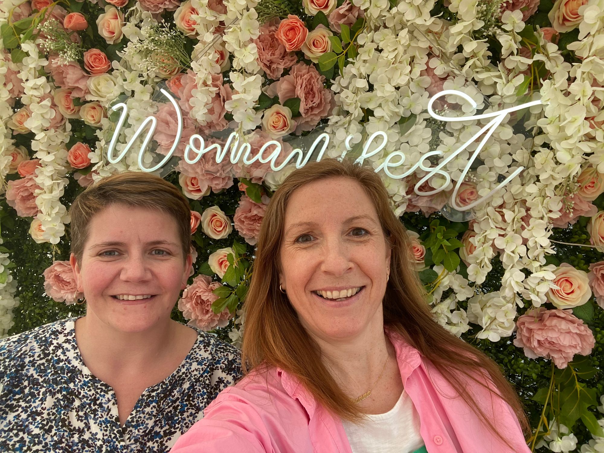 Maggie and Joanne standing in front of a flower wall with the Womanifest neon sign behind