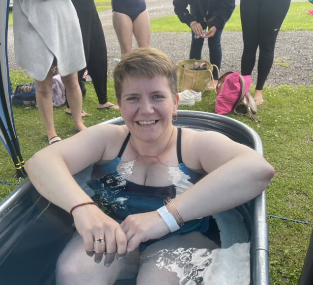 Maggie smiling in swimsuit in ice-cold water in a steel bathtub on grass as other participants wait their turn.
