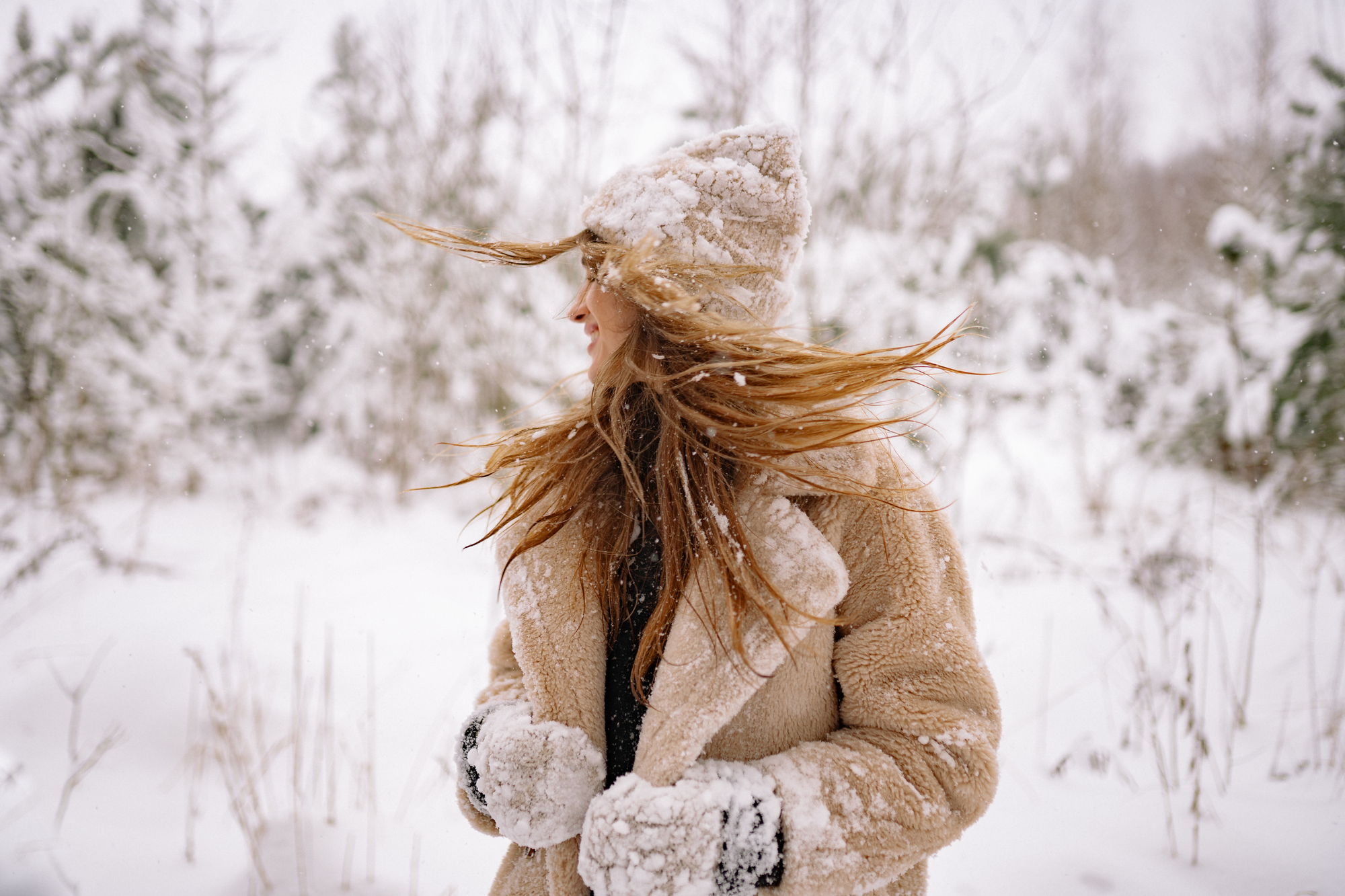 Women in snow surrounded by trees, in fluffy cream coat , shaking the snow from her head 